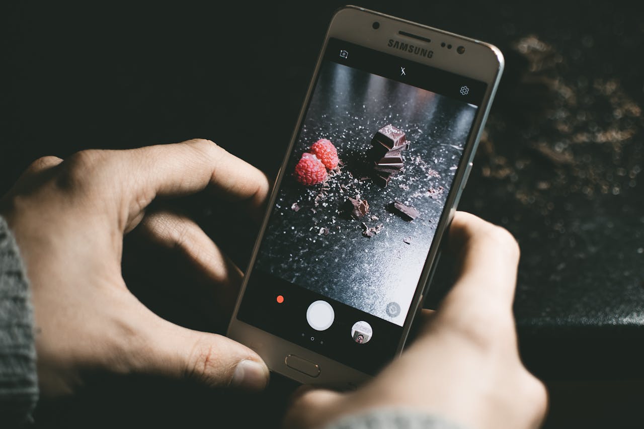 Close-up of a smartphone photographing chocolate pieces and raspberries on a dark surface.
